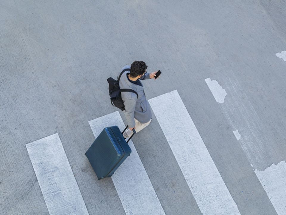 A man in a crosswalk pulling a suitcase and looking at his phone.