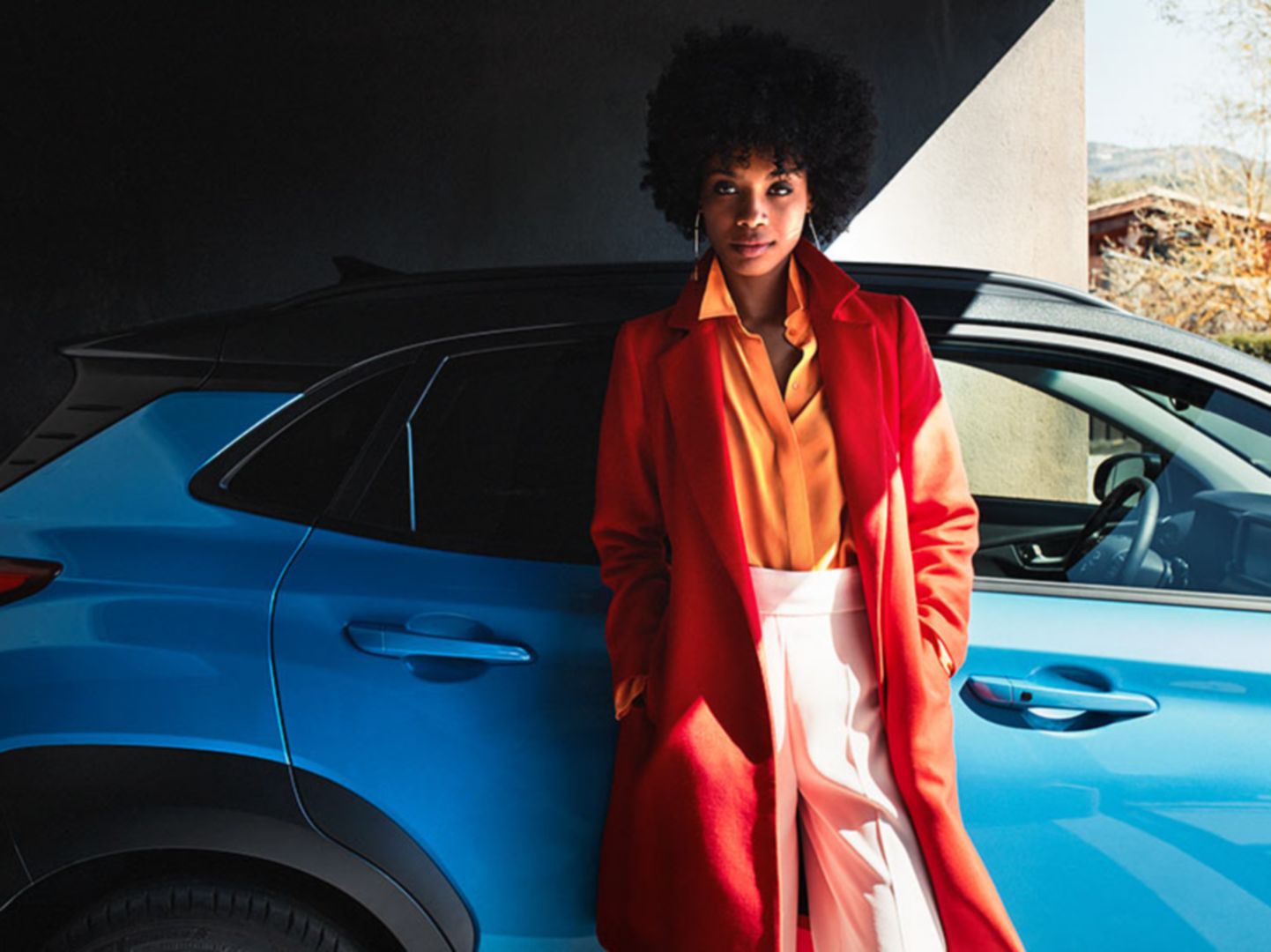 A woman in a long red coat, leaning against the passenger door of her Hyundai KONA Hybrid.